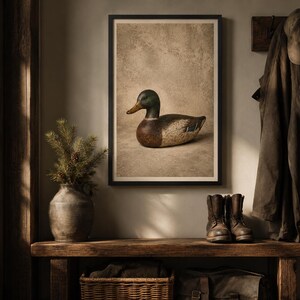 Pode incluir: Uma impress&atilde;o art&iacute;stica emoldurada apresentando um chamariz de pato-real com cabe&ccedil;a verde, bico amarelo e corpo castanho e branco, exibida numa sala com um banco de madeira, um vaso de cer&acirc;mica com ramos de pinheiro, um casaco, um bon&eacute; e botas de couro.