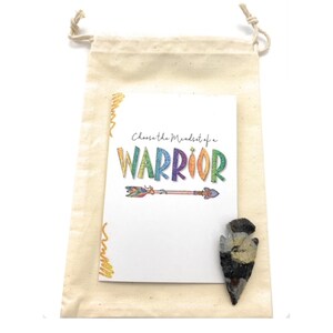 May include: A natural canvas drawstring bag containing a card with the text "Choose the Mindset of a WARRIOR" and a colorful arrow illustration. A small, dark arrowhead-shaped stone rests beside the card.