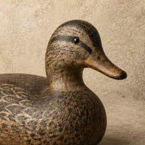 Pode incluir: Um chamariz de pato em madeira esculpida com um padr&atilde;o de plumagem texturizado em castanho e preto. A figura tem uma cabe&ccedil;a castanho-escura com uma faixa preta no olho, um bico castanho-claro e um corpo arredondado com penas detalhadas.