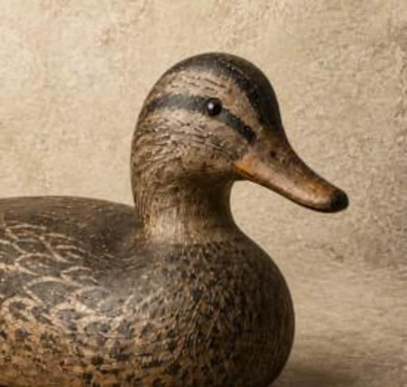 Pode incluir: Um chamariz de pato em madeira esculpida com um padr&atilde;o de plumagem texturizado em castanho e preto. A figura tem uma cabe&ccedil;a castanho-escura com uma faixa preta no olho, um bico castanho-claro e um corpo arredondado com penas detalhadas.