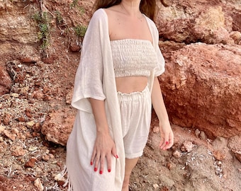 Könnte beinhalten: Ein cremefarbenes zweiteiliges Set bestehend aus einem trägerlosen, gesmokten Schlauchtop und passenden Shorts mit hoher Taille und elastischem Bund, kombiniert mit einem langen, vorne offenen Kimono mit Fransen am unteren Saum.