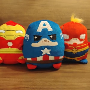 May include: Three plush toys in the shape of superheroes. The Iron Man toy is red and yellow, Captain America is blue with a white star, and Captain Marvel is red and yellow. They are on a wooden surface.