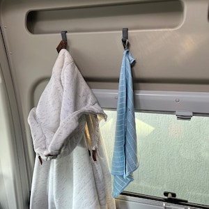 Op de afbeelding: Een witte hoodie en een blauw-wit gestreepte handdoek hangen aan haken in een voertuig. De hoodie heeft een gespikkeld patroon en bruine lederen accenten. De handdoek is opgevouwen.