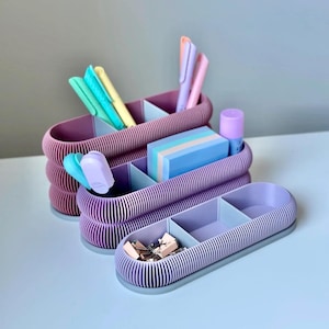 Könnte beinhalten: Drei pastellfarbene Schreibtisch-Organizer mit mehreren Fächern. Die Organizer sind mit Stiften, Büroklammern und Haftnotizen gefüllt. Die Organizer sind in Lila-, Lavendel- und Blautönen gehalten. Das Design weist eine gerippte Textur auf.
