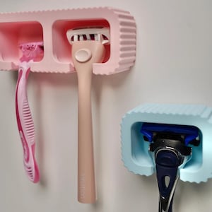May include: A light pink wall-mounted holder with two slots containing a pink razor and a peach-coloured Intuition razor, alongside a separate light blue wall-mounted holder containing a dark blue and silver razor, all featuring a ribbed exterior design.