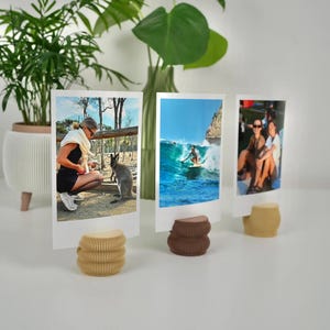 May include: Three photos displayed on decorative paper stands. The photos show a person with a kangaroo, a surfer on a wave, and two people. The stands are in shades of beige and brown, and the background includes plants.