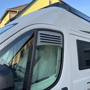 Könnte beinhalten: Weißer Campingbus mit schwarzer Verkleidung und einem Seitenfenster. Das Fenster hat eine Lüftung und einen schwarzen Rahmen. Der Van ist an einem sonnigen Tag draußen geparkt. Das Wort "SUM" ist auf der Seite des Vans zu sehen.