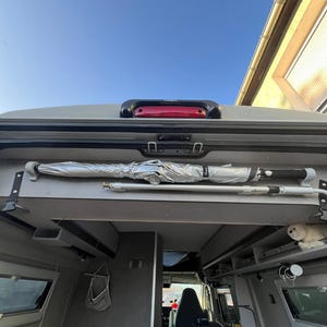 Puede incluir: Un paraguas plateado con mango negro está montado horizontalmente en el techo interior de una furgoneta camper. Debajo del paraguas, un poste telescópico blanco está asegurado. El interior de la furgoneta tiene paredes grises, ventanas y una cama.