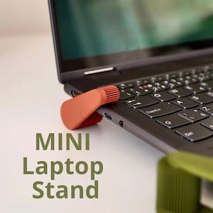Può includere: Un mini supporto per laptop arancione bruciato sostiene un laptop nero. Il supporto è angolato per sollevare il laptop. Il testo "MINI Laptop Stand" è stampato in verde. La tastiera è nera con tasti bianchi.