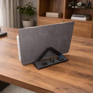 Può includere: Un tablet grigio appoggiato su un supporto nero con una penna bianca. Il supporto ha un supporto triangolare e una piccola base con pulsanti e una chiavetta USB blu. Su una scrivania di legno.