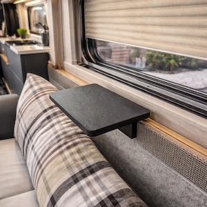 Può includere: Interno di un camper con un piccolo tavolo rettangolare nero attaccato al muro. Sono visibili un cuscino a quadri e una finestra con una tenda beige. Il camper ha pannelli in legno chiaro e una zona cucina.