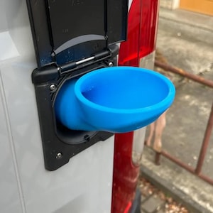 Puede incluir: Un puerto de llenado de agua azul brillante, en forma de embudo, montado en una superficie blanca de un vehículo. El puerto está situado dentro de una carcasa negra con una tapa con bisagras. La luz trasera roja del vehículo es visible en el fondo.
