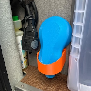 Könnte beinhalten: Ein blau-oranges 3D-gedrucktes Objekt, möglicherweise ein Halter oder Organizer, ist an einer Wand montiert. Ein schwarzes Netzkabel mit Stecker ist sichtbar, zusammen mit einer Flüssigkeitsflasche. Das Objekt befindet sich in einem Fach.