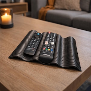 May include: A black, wavy-shaped remote control holder with two remotes on a wooden table. One remote is a Fire TV remote, and the other is a Samsung remote. A lit candle in a glass holder is in the background.