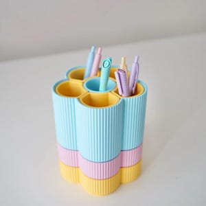 Könnte beinhalten: Ein pastellfarbener Schreibtisch-Organizer mit sechs Fächern, in denen jeweils ein Stift untergebracht ist. Der Organizer ist zylindrisch mit vertikalen Rippendetails. Die Farben sind Hellblau, Rosa und Gelb.