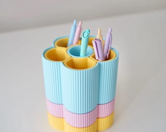 Może przedstawiać: Pastelowy organizer biurowy z sześcioma przegródkami, z których każda mieści długopis. Organizer jest cylindryczny z pionowymi żebrowanymi detalami. Kolory to jasnoniebieski, różowy i żółty.