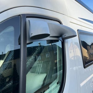 Könnte beinhalten: Nahaufnahme eines weißen Wohnmobils mit einem grauen Luftventilationssystem, das über dem Fenster montiert ist. Die Lüftung hat zwei Öffnungen mit einem Mesh-Gitter. Das Fenster spiegelt das Innere wider, einschließlich eines weißen Kissens. Die Seite des Vans ist sichtbar.