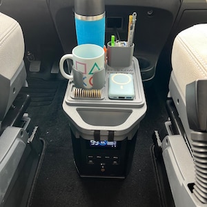 May include: A black and grey portable power station with a tray holding a blue tumbler, a mug with colourful geometric shapes, a pen holder, and a wireless charger. The power station has a digital display.