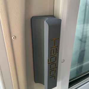 May include: A gray door handle with the word "Hägdor" in gold lettering. The handle is mounted on a beige wall next to a white door frame and a glass window.