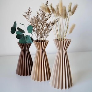 Può includere: Tre vasi decorativi nei toni del marrone e del beige, ciascuno con piante essiccate. I vasi hanno un design a pieghe e sono disposti su una superficie bianca. Il vaso a sinistra è marrone scuro, il vaso centrale è beige e il vaso a destra è beige chiaro.