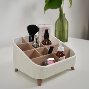 Pode incluir: Um organizador de maquilhagem branco e bege com vários compartimentos, contendo vários produtos de beleza. O organizador tem um exterior texturizado e assenta em pequenas pernas de madeira. Uma escova preta, frascos e tubos são visíveis.