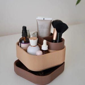 Op de afbeelding: Een tweekleurige, geribbelde cosmetica-organizer in bruin en beige, gevuld met schoonheidsproducten. De organizer bevat flessen, tubes, borstels en andere items. De bovenste laag is beige en de onderste laag is bruin.