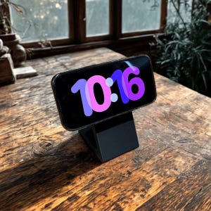 Könnte beinhalten: Ein schwarzes Smartphone, das die Uhrzeit 10:16 in Pink und Lila anzeigt, ruht auf einem schwarzen Ständer. Der Ständer steht auf einem Holztisch mit natürlicher Maserung. Der Hintergrund umfasst ein Fenster und Grün.