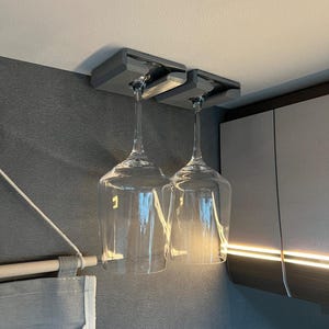 Op de afbeelding: Twee heldere wijnglazen hangen ondersteboven aan een grijze, aan het plafond gemonteerde houder. De houder is gemaakt van een solide, rechthoekig materiaal. De glazen hangen boven een donkergrijze muur, met een glimp van een kast op de achtergrond.