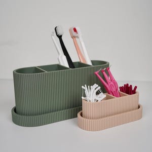 Pode incluir: Dois recipientes ovais e canelados, um verde sálvia e outro rosa claro, contêm artigos de higiene dentária. O recipiente maior contém três escovas de dentes com cabos brancos, pretos e pêssego. O recipiente menor contém palitos de fio dental e escovas interdentais.