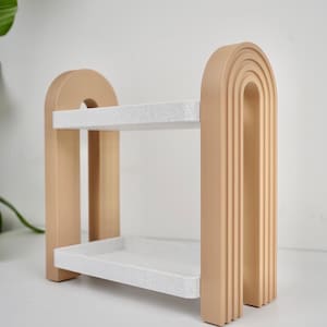 Può includere: Una mensola decorativa a due livelli dal design moderno. La struttura è di colore beige chiaro, con una forma ad arco arrotondata. I ripiani sono rettangolari e presentano un motivo bianco e grigio maculato. La mensola è adatta per esporre piccoli oggetti.