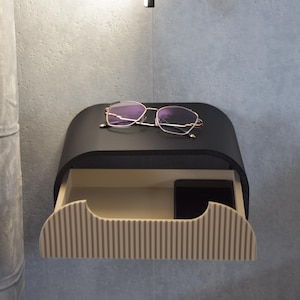 Peut inclure: Une table de chevet murale noire avec un tiroir ouvert, contenant une paire de lunettes et un smartphone. Le tiroir révèle un intérieur clair et un panneau avant à rayures verticales. La table est fixée sur un mur gris.