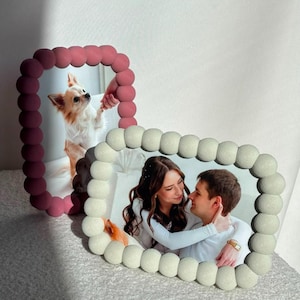 Op de afbeelding: Twee rechthoekige fotolijsten met een textuur van bolletjes langs de rand. De ene lijst is donkerroze en bevat een foto van een kleine hond. De andere lijst is crèmekleurig en bevat een foto van een koppel dat elkaar omhelst.