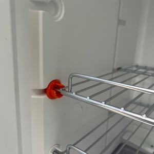 Può includere: Vista interna di un frigorifero bianco con ripiani in filo metallico. Una clip di plastica arancione fissa un ripiano. L'interno del frigorifero è pulito e ben illuminato, con un focus sul sistema di scaffalature e sui suoi componenti.