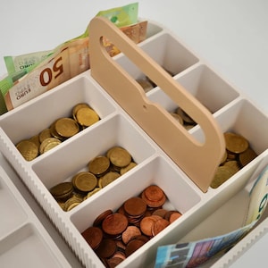 Könnte beinhalten: Eine weiße Kunststoff-Geldkassette mit mehreren Fächern zur Organisation von Bargeld. Die Kassette verfügt über einen hellen Mittelgriff und enthält verschiedene goldfarbene Münzen sowie Euro-Banknoten, darunter ein sichtbarer 50-Euro-Schein.