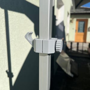Peut inclure: Une pince en plastique gris clair avec un crochet intégré et un bouton de serrage circulaire, fixée solidement sur un poteau vertical en métal argenté de section carrée. L'élément présente une conception en deux parties avec une molette.