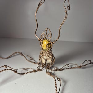 Könnte beinhalten: Handgefertigte Drahtskulptur mit einem zentralen gelb-weißen Stein, ausgestreckten Armen und mehreren Drahtbeinen. Die Skulptur besteht aus kupferfarbenem Draht mit filigranen Details und einem einzigartigen, künstlerischen Design.