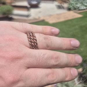 May include: A copper-colored ring with a braided design. The ring is worn on a finger, showcasing its intricate woven pattern. The ring's warm hue contrasts with the skin tone, highlighting the craftsmanship of the jewelry.