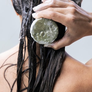 Op de afbeelding: De rug en het haar van een persoon zijn bedekt met shampooschuim. Een ronde, groene zeep met het woord "TONS" wordt vastgehouden in de hand van de persoon, die zich op de achterkant van hun hoofd bevindt. Het haar is donker en nat.