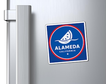 May include: A square magnet with a blue background and a red border. The magnet features the text "ALAMEDA CALIFORNIA" and a white graphic of a watermelon slice. The magnet is attached to a silver refrigerator door.