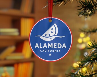 May include: A round Christmas ornament with a blue background and a white and red border. The ornament features a white watermelon slice design and the text "ALAMEDA CALIFORNIA" in white. A red ribbon is attached for hanging.