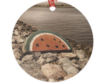 May include: A round ornament featuring a painted watermelon slice on a rocky shoreline with a body of water in the background. The watermelon has a red interior, black seeds, and a green rind. A red ribbon is attached for hanging.