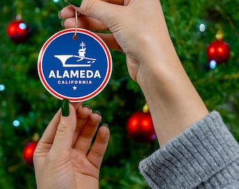 May include: A round Christmas ornament with a blue background and a white graphic of a ship. The ornament has the words "ALAMEDA CALIFORNIA" in white text. A gold string is attached for hanging.