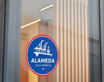 May include: A round sticker with a blue background and a red border. The sticker features a white illustration of a bridge and a ship, with the word "ALAMEDA" and "CALIFORNIA" in white text. A white star is at the bottom.