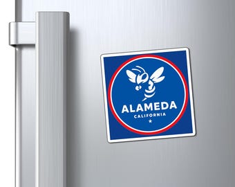 May include: A square magnet with a blue background, a red circle, and a white border. The magnet features a white bee graphic and the text "ALAMEDA CALIFORNIA". The magnet is attached to a silver refrigerator door.