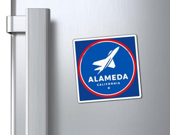May include: A square magnet with a blue background, a red circle, and white text that reads "Alameda California". A white jet graphic is inside the circle. The magnet is attached to a silver refrigerator door.