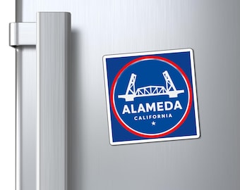 May include: A square magnet with a blue background and a red circular border. The magnet features a white illustration of a bridge and the text "ALAMEDA CALIFORNIA" in white. A star is at the bottom.