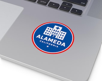 May include: A round sticker featuring the text "ALAMEDA CALIFORNIA" in white, with a star below, and a graphic of a building with a cross on top. The design is on a blue background with a red border.