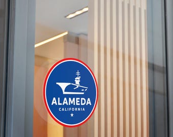 May include: A round sticker with a blue background and a white illustration of a ship. The sticker has a red border and the words "ALAMEDA CALIFORNIA" in white text. A white star is at the bottom.