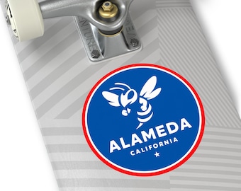 May include: A sticker featuring a blue circle with a white bee graphic and the word "ALAMEDA" in white text. The sticker also includes "CALIFORNIA" and a star. The sticker is on a skateboard.