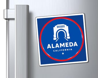 May include: A square magnet featuring a blue background with a white illustration of a bridge and the text "ALAMEDA CALIFORNIA" in white. A red circle surrounds the bridge illustration. The magnet is attached to a silver refrigerator.